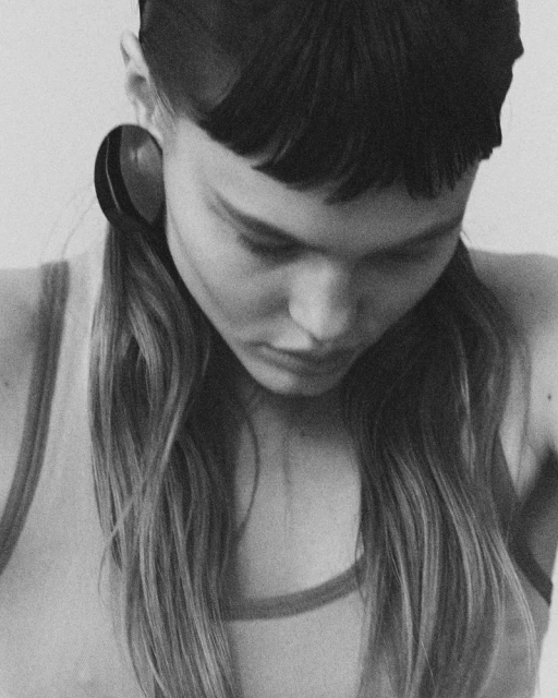 Monochrome portrait of a young adult female model with blunt bangs, long wavy hair, wearing large hoop earrings and a tank top.