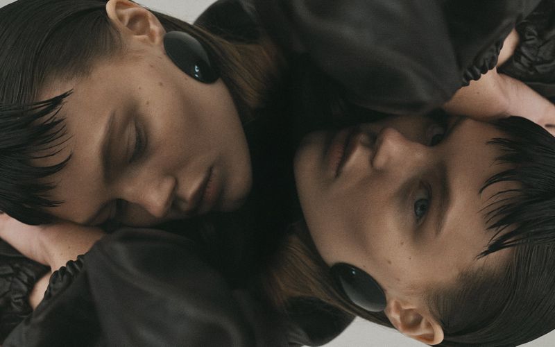 Close-up portrait of two young women with similar features, dark hair, and black disc earrings, lying close together in soft lighting.