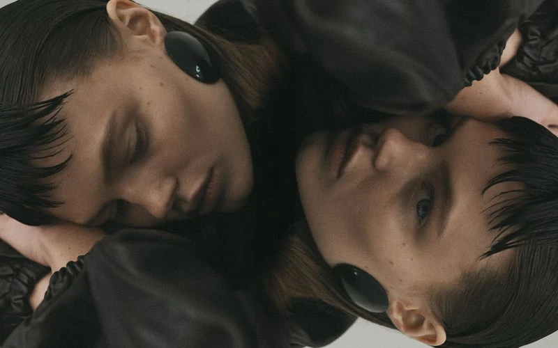 Close-up portrait of two young women with similar features, dark hair, and black disc earrings, lying close together in soft lighting.