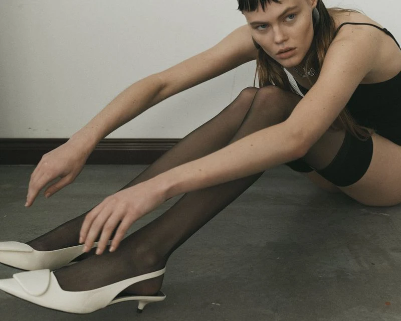 Young woman poses indoors sitting on floor in dark top, black stockings, white slingback heels, and grey stone background. 