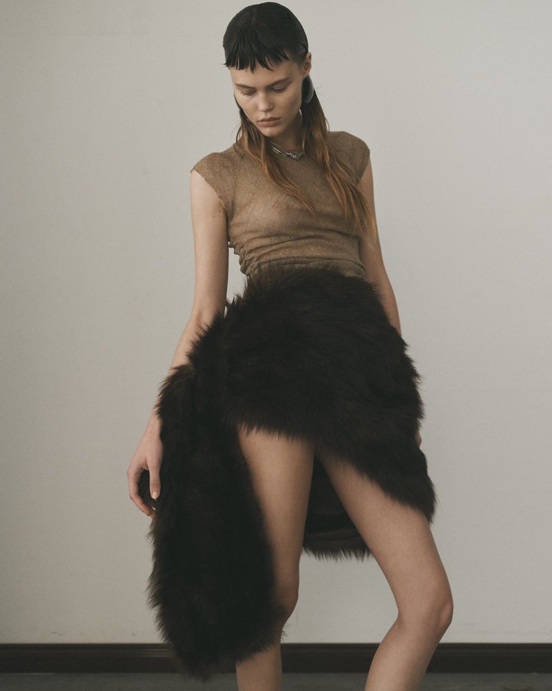 Woman in a tan top and dark brown faux fur skirt, posing against a light wall with dark floor, fashion studio shot.