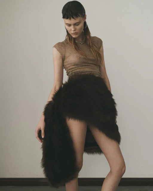 Woman in a tan top and dark brown faux fur skirt, posing against a light wall with dark floor, fashion studio shot.