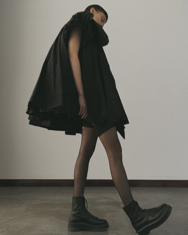 A person in a black, oversized, draped coat and black boots poses in front of a neutral wall in a fashion-forward studio shot.