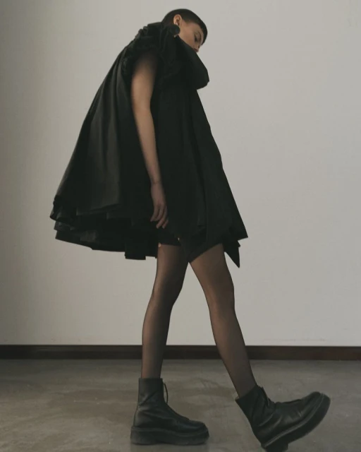 A person in a black, oversized, draped coat and black boots poses in front of a neutral wall in a fashion-forward studio shot.
