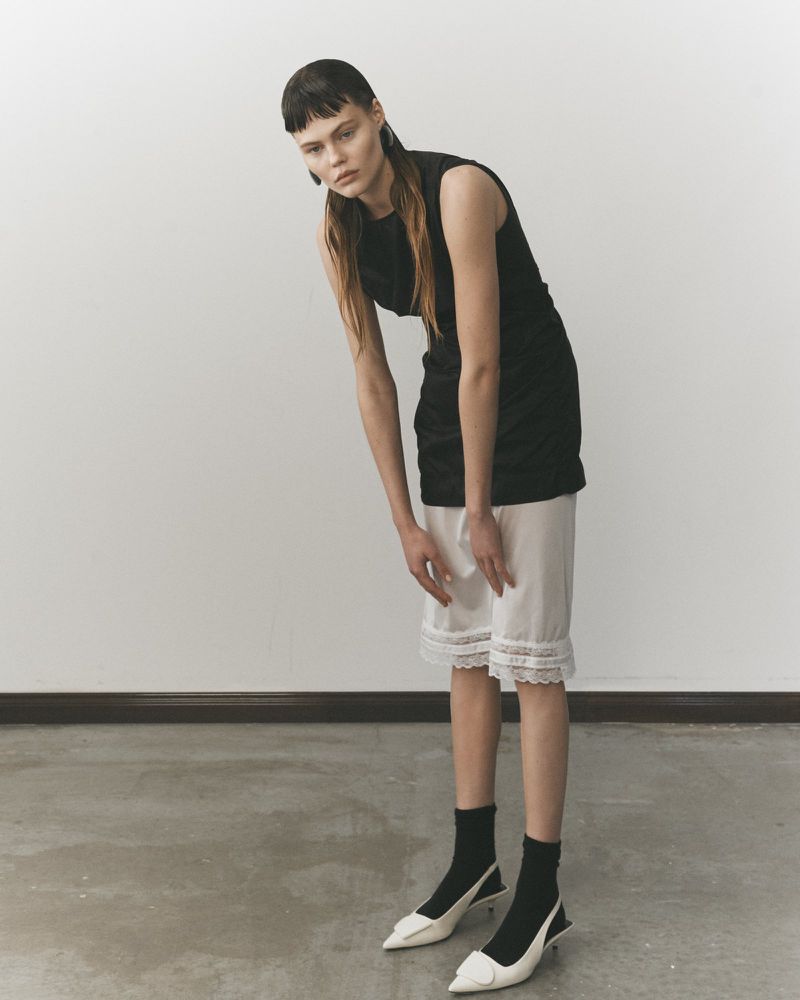 A slender young woman stands in a minimalist photo studio, wearing a black top, white lace-trimmed shorts, black socks, and white heels.