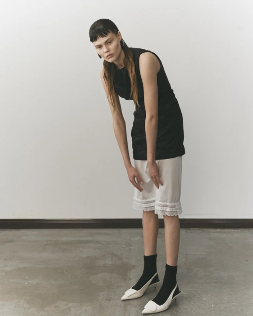 A slender young woman stands in a minimalist photo studio, wearing a black top, white lace-trimmed shorts, black socks, and white heels.