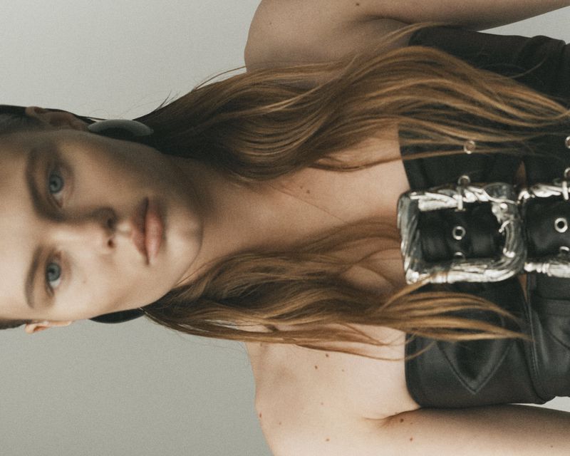 Close-up studio portrait of a young adult woman with long wavy auburn hair wearing a black corset top with silver buckles.