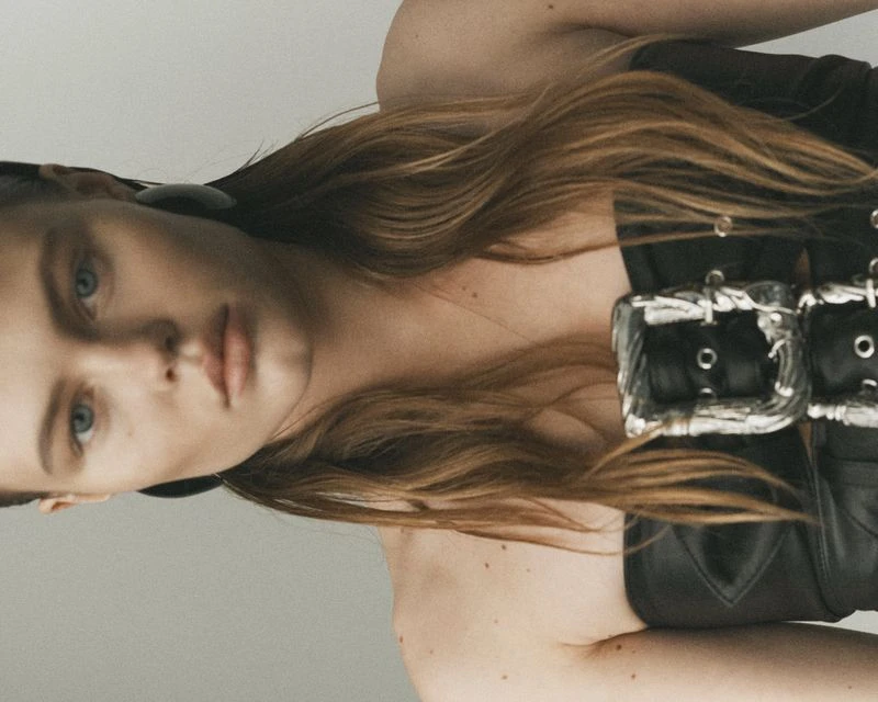 Close-up studio portrait of a young adult woman with long wavy auburn hair wearing a black corset top with silver buckles.