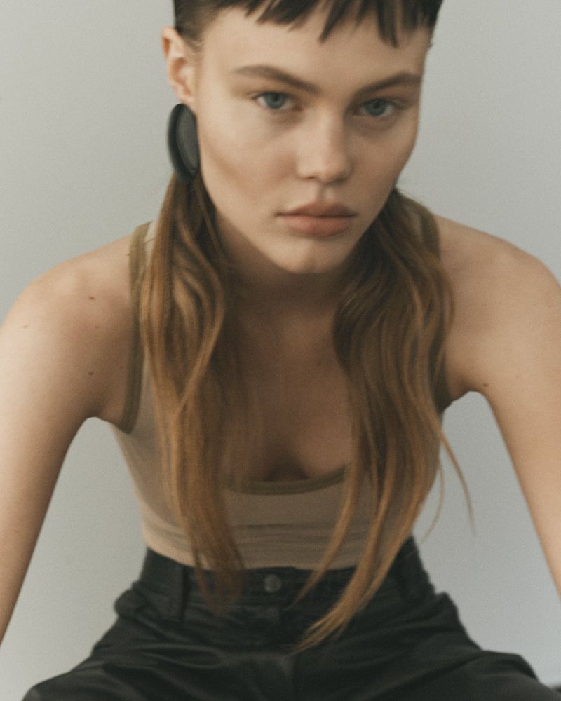 Close-up of a young fair-skinned woman with light brown hair and black hoop earrings wearing a tan tank top and black pants.