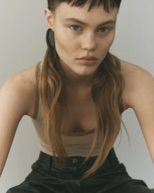 Close-up of a young fair-skinned woman with light brown hair and black hoop earrings wearing a tan tank top and black pants.