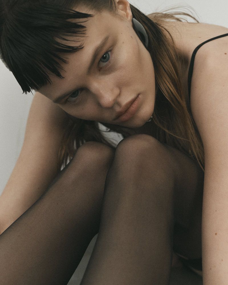 A young woman with light skin and dark hair poses indoors with sheer black pantyhose and a black tank top in soft, neutral lighting.