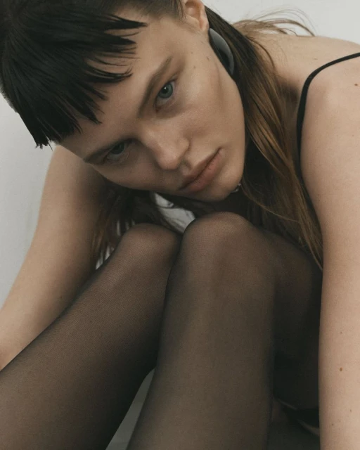 A young woman with light skin and dark hair poses indoors with sheer black pantyhose and a black tank top in soft, neutral lighting.