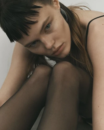 A young woman with light skin and dark hair poses indoors with sheer black pantyhose and a black tank top in soft, neutral lighting.