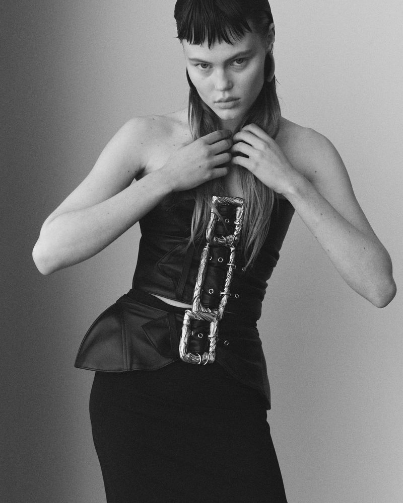 Black and white studio portrait of a woman with a severe hairstyle wearing a structured corset and a pencil skirt.