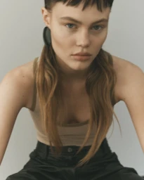 Model Kiki Jewel leaning forward in a beige tank top and black trousers against a neutral studio backdrop.