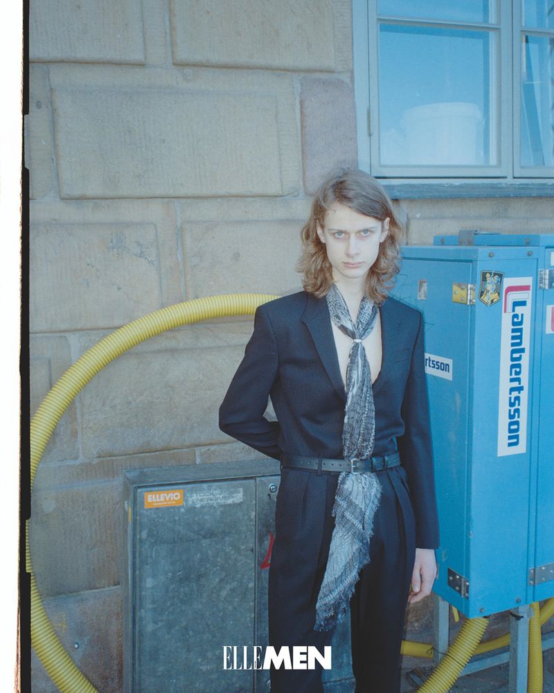 Editorial fashion shot of a long-haired man in a black suit and patterned scarf, posing outdoors against a stone wall with utility boxes.