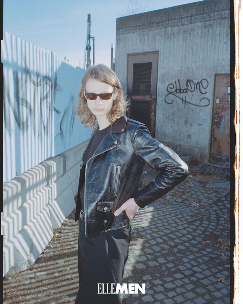 Adult standing on brick sidewalk wearing sunglasses and leather jacket in front of a concrete building with graffiti.