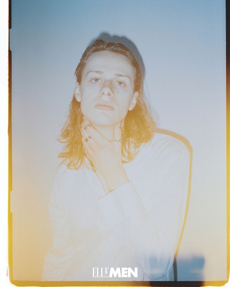 Indoor portrait of a young adult with long hair wearing a white shirt and several rings, holding a hand to their neck in soft light.