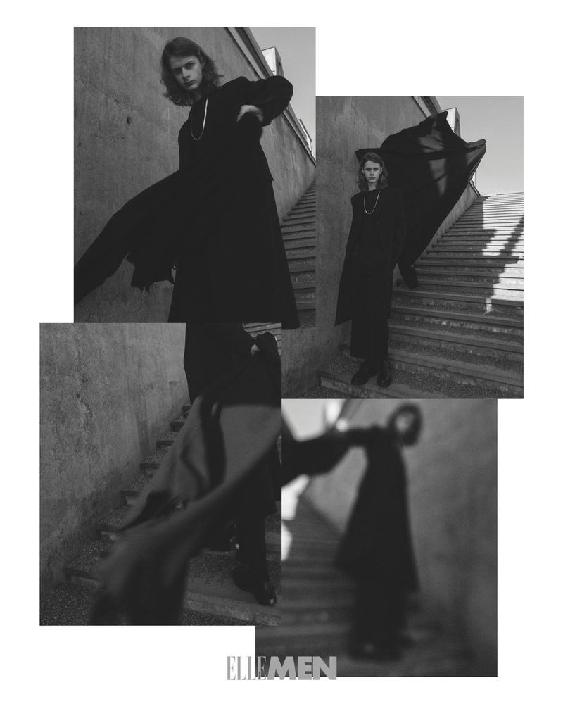 Monochrome fashion portrait of a person in a long black coat and necklace, standing on outdoor steps, in an editorial style.