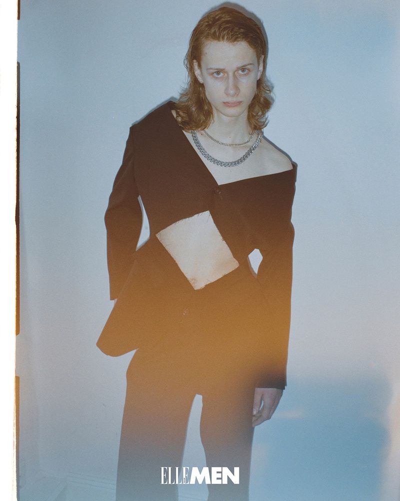 A slender, light-skinned male model with reddish-brown hair in a black deconstructed blazer and matching pants against a gradient light-blue backdrop.
