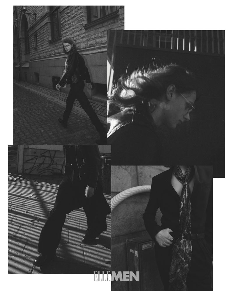 Monochrome fashion shot of a slender adult male model wearing a leather jacket and suit, posing outdoors on a city street with ELLE Men logo.