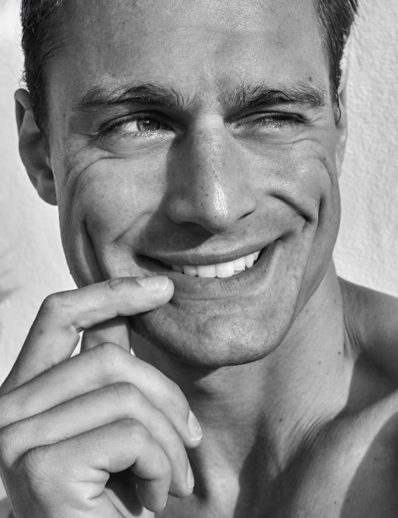 Black and white portrait of a smiling man with a hand near his mouth, set against a neutral background, capturing a moment of happiness.