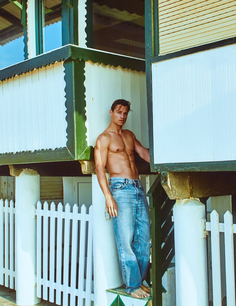 Shirtless man in jeans stands near an elevated beach house with a white picket fence, captured in bright daylight with a medium shot.