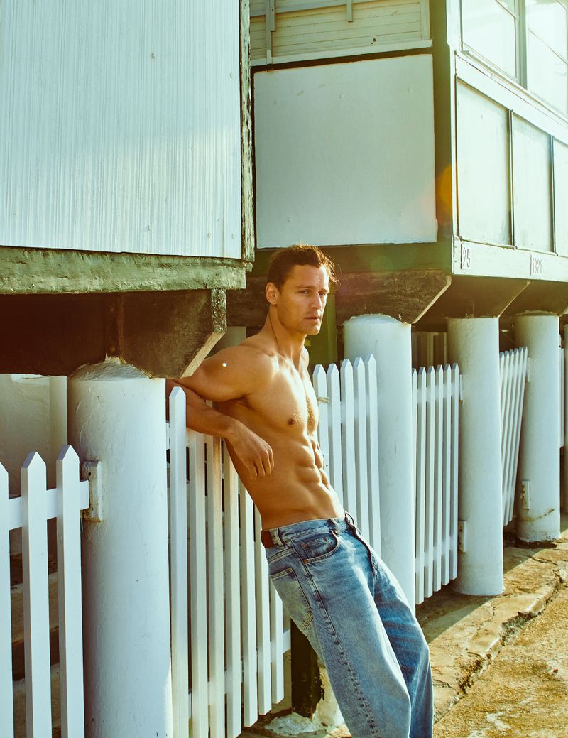 Shirtless male model in jeans leaning against a white fence outside a building. He has an athletic build and is in bright sunlight.