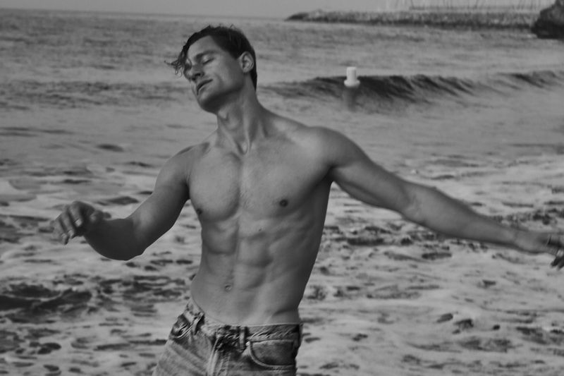 Black and white portrait of a muscular adult male standing shirtless on a beach with the ocean and sky in the background.