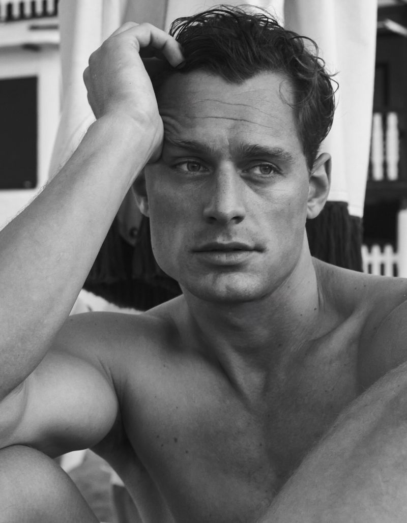 Black and white close-up portrait of a shirtless man running his hand through his wavy hair against a blurred outdoor backdrop.