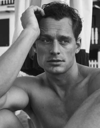 Black and white close-up portrait of a shirtless man running his hand through his wavy hair against a blurred outdoor backdrop.