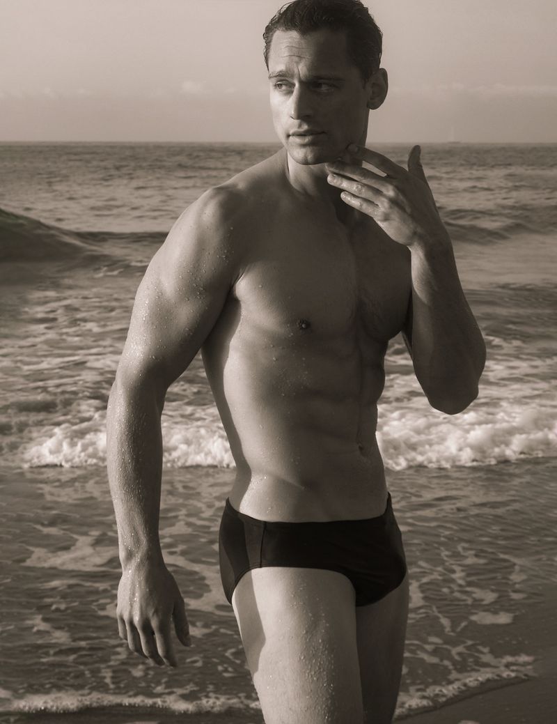 Monochrome portrait of a muscular adult male in swimwear standing on a beach with gentle waves and a cloudy sky in the background.