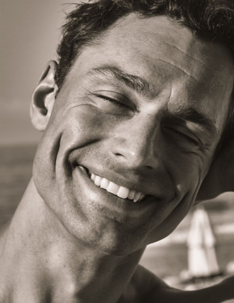 Close-up of a smiling adult man with closed eyes in a portrait framed against a neutral outdoor setting with out-of-focus background.