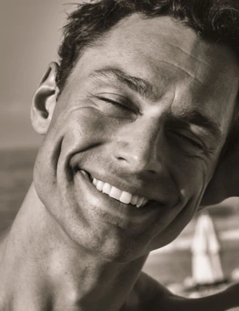 Close-up of a smiling adult man with closed eyes in a portrait framed against a neutral outdoor setting with out-of-focus background.