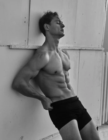 Shirtless athletic man in black shorts leaning back against a paneled wall in a black and white portrait.