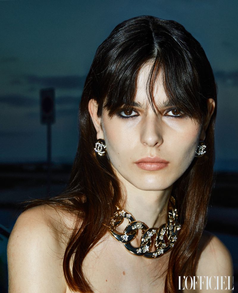 Close-up of a brunette woman in earrings and a gold necklace, set against an outdoor background with a muted dusk sky.