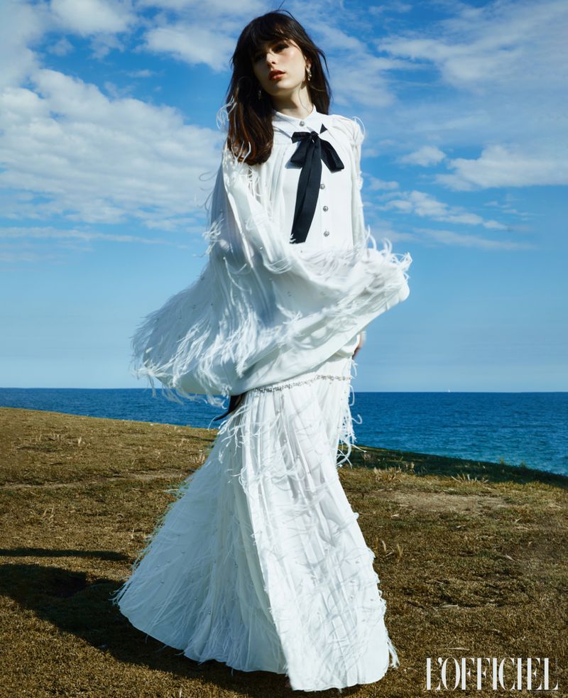 Fashion editorial image of a woman in a white dress with a black bow standing on a grassy hill overlooking the ocean under a blue sky.