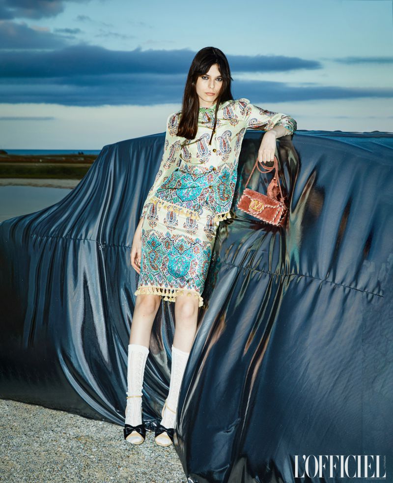 A fashionable woman in a paisley print top and skirt poses on a car covered with a black tarp in a landscape setting, holding a purse.