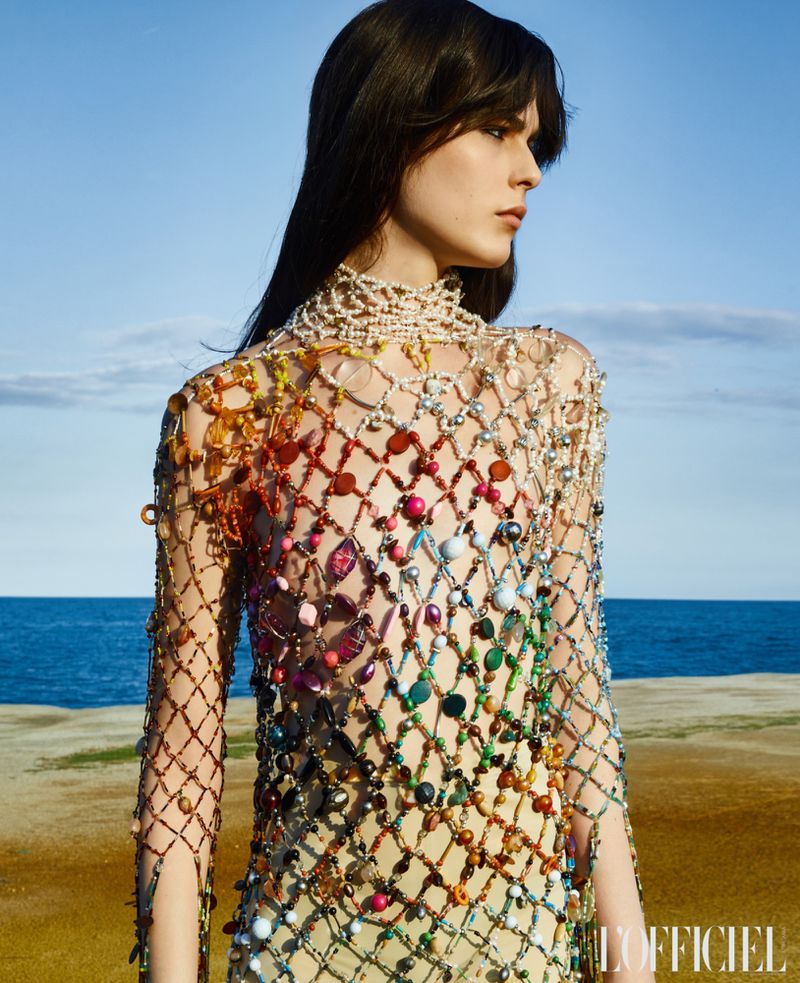 Brunette woman poses in a colorful beaded dress outdoors, against a backdrop of blue sky, tan land, and ocean in a fashion editorial.