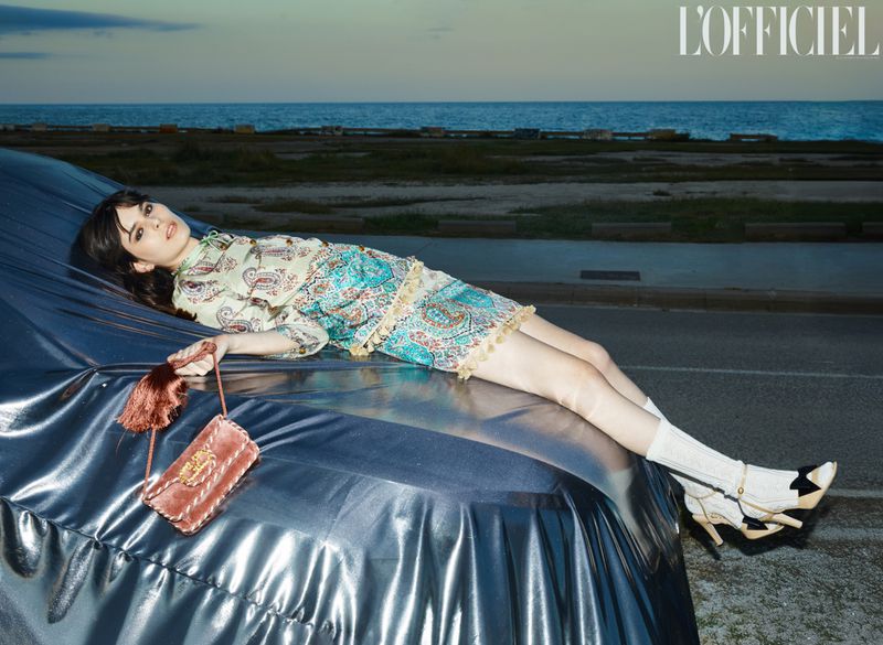 Young woman poses on a vintage car under a silver tarp near the ocean, wearing a patterned dress, white socks, and carrying a purse.