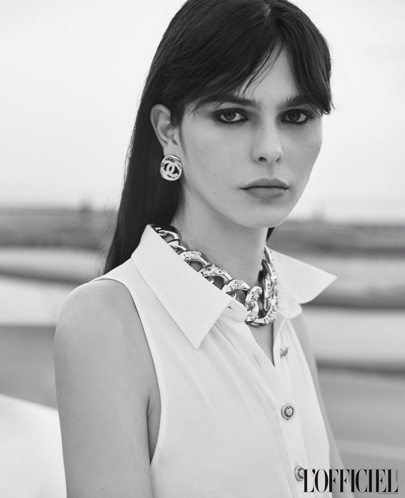 Monochrome fashion portrait of a model wearing a white sleeveless shirt, chunky chain necklace, and logo earrings.
