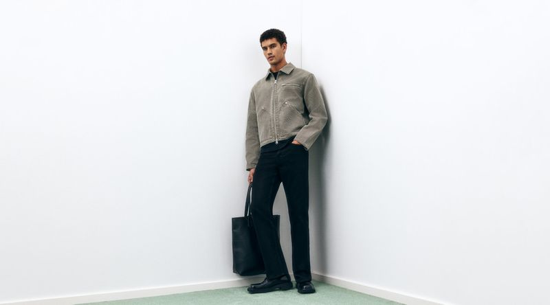 A young adult man poses in a studio, wearing an olive-green jacket, black pants, and black shoes, holding a black tote bag.