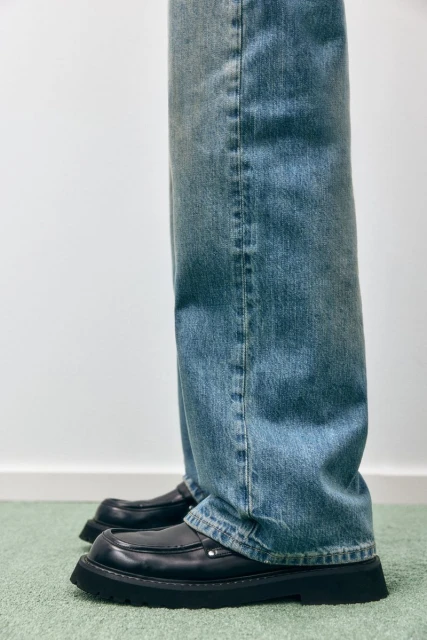 Close-up of jeans and black leather loafers. The person is standing on a green carpet with a plain white wall in the background.