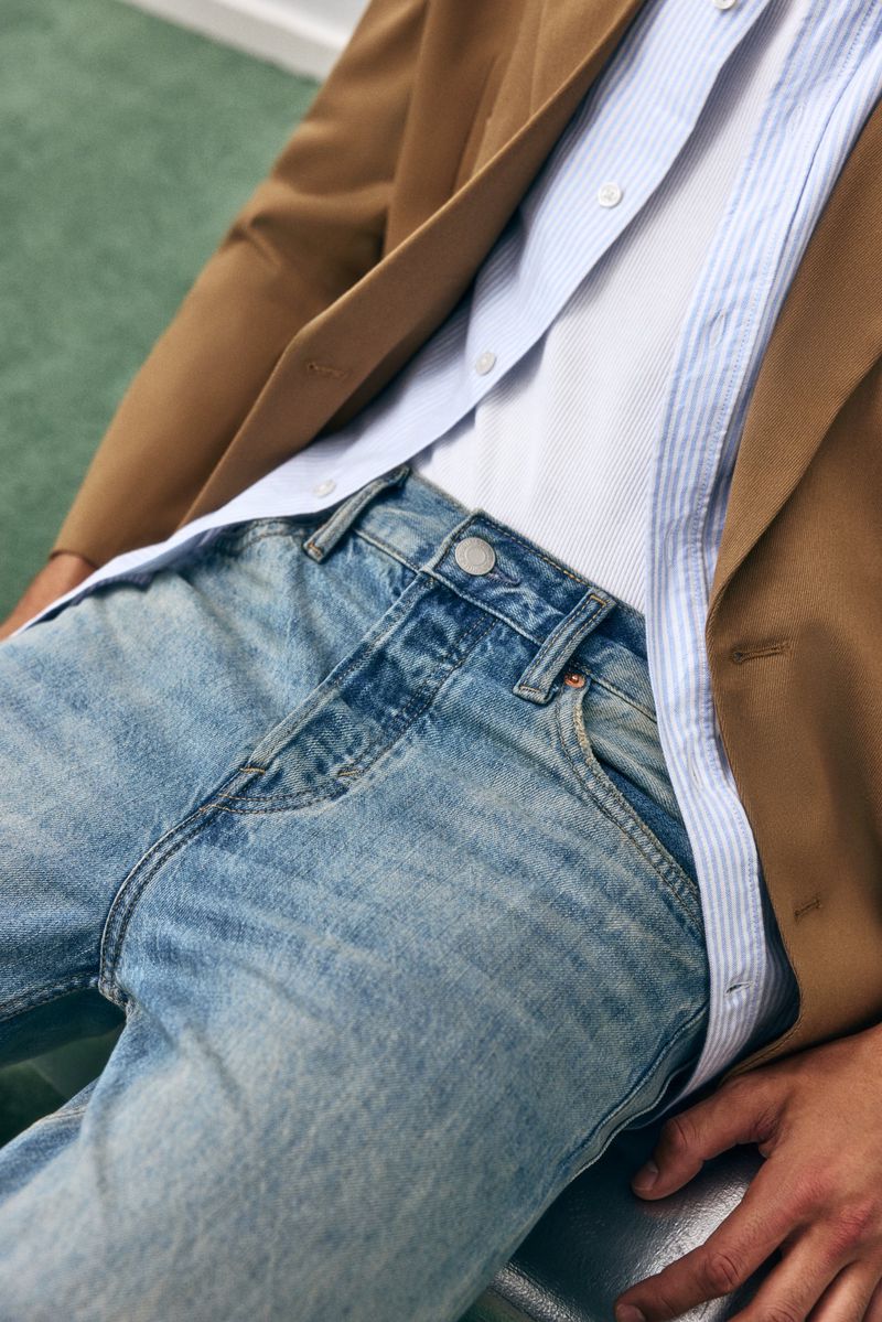 Close-up studio shot of a fair-skinned adult man wearing light-wash jeans, a white t-shirt, blue dress shirt, and tan blazer.