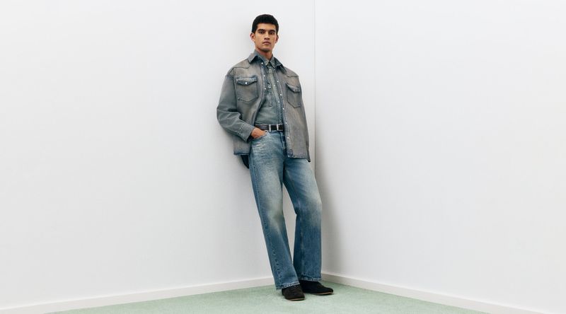 Fashion shot of a man leaning against a wall wearing a denim shirt and jeans, showcasing a monochromatic, denim-on-denim fashion style.