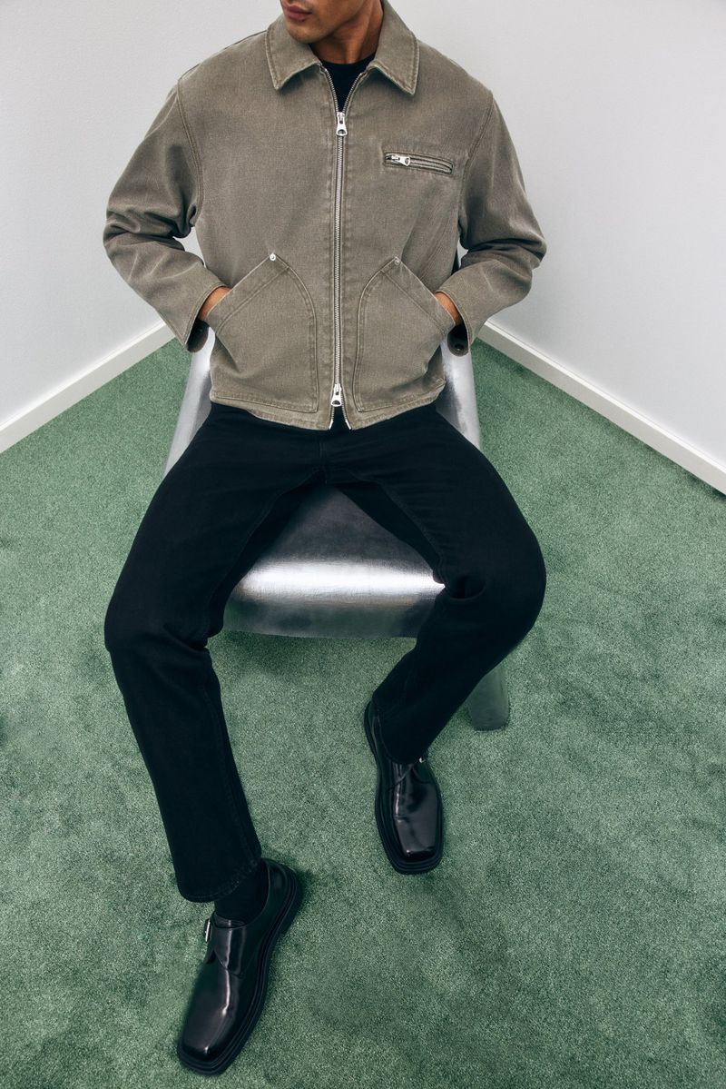 A seated adult male poses wearing a grey-brown jacket, black pants, and black shoes against a white wall and green carpet.