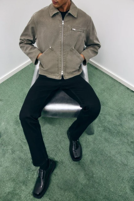A seated adult male poses wearing a grey-brown jacket, black pants, and black shoes against a white wall and green carpet.