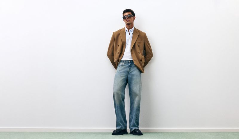 Model Bodhi Heeck wearing a camel blazer, white shirt, light wash jeans, and sunglasses against a minimal white wall.
