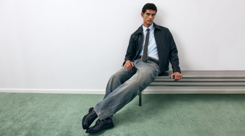 Male model in jacket, striped shirt, tie, and jeans sits on a gray bench against a white wall and green carpet, casual fashion studio shot.