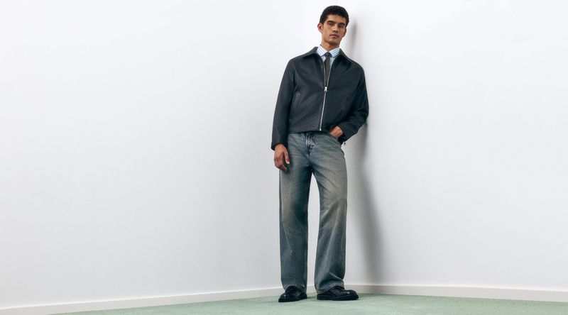 Model Bodhi Heeck leans against a white wall wearing a dark zip-up jacket and wide-leg faded denim jeans.